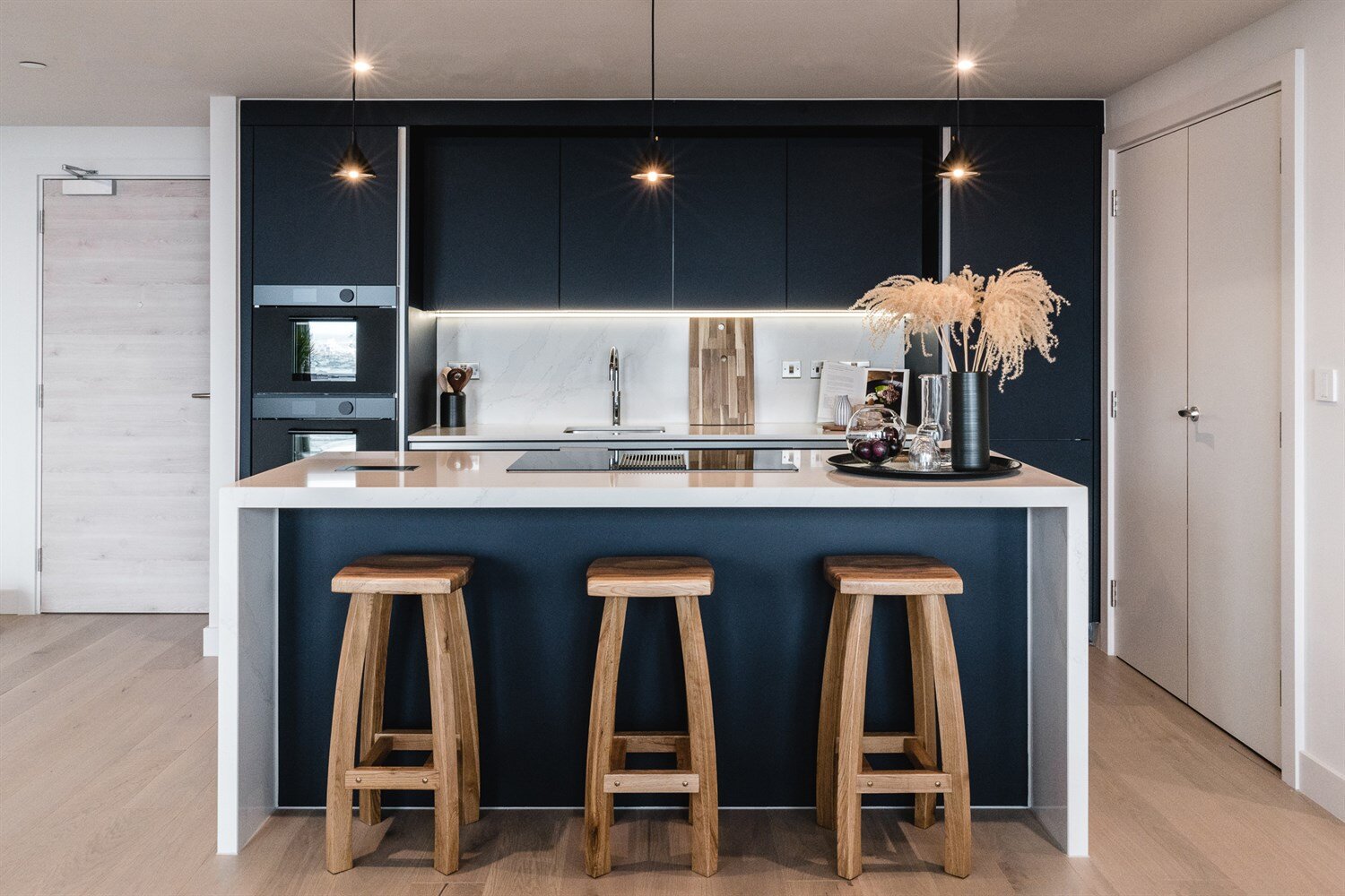 Navy kitchen with integrated ovens, a marble waterfall island and three oak stools