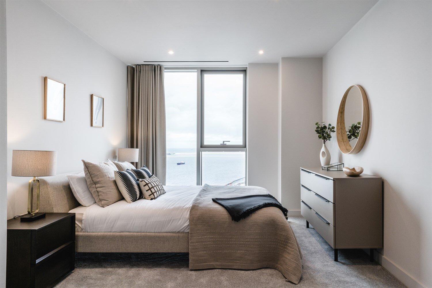 Bedroom with a linen headboard, a brass lamp and a tall window onto open water