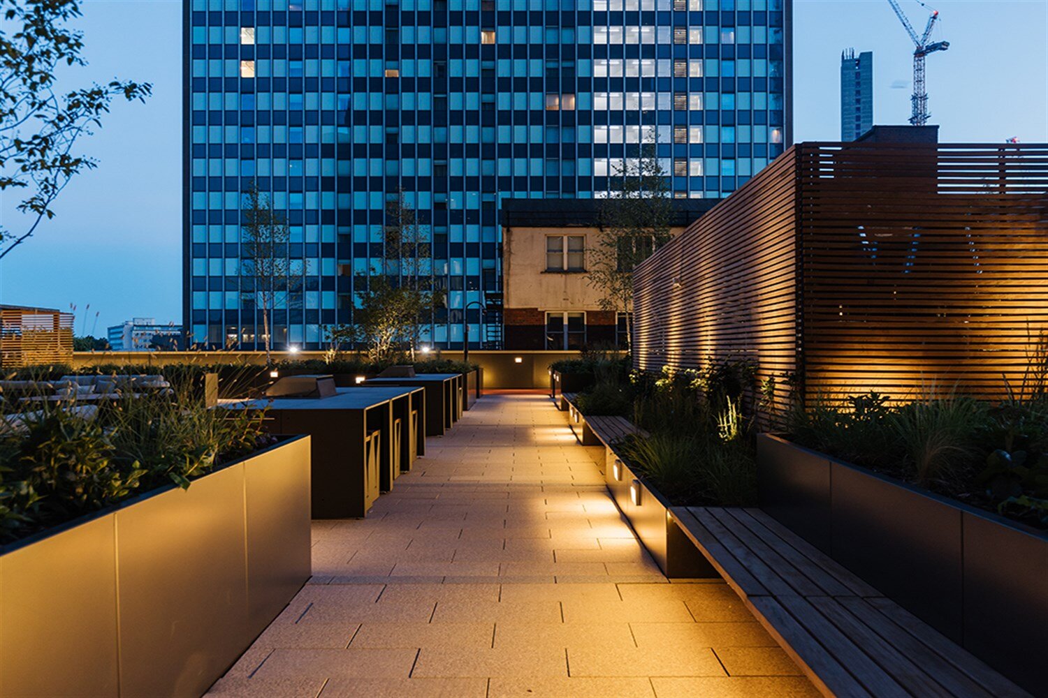 Roof terrace at dusk with lit planters, timber screens and towers beyond, at The Mercian, Birmingham