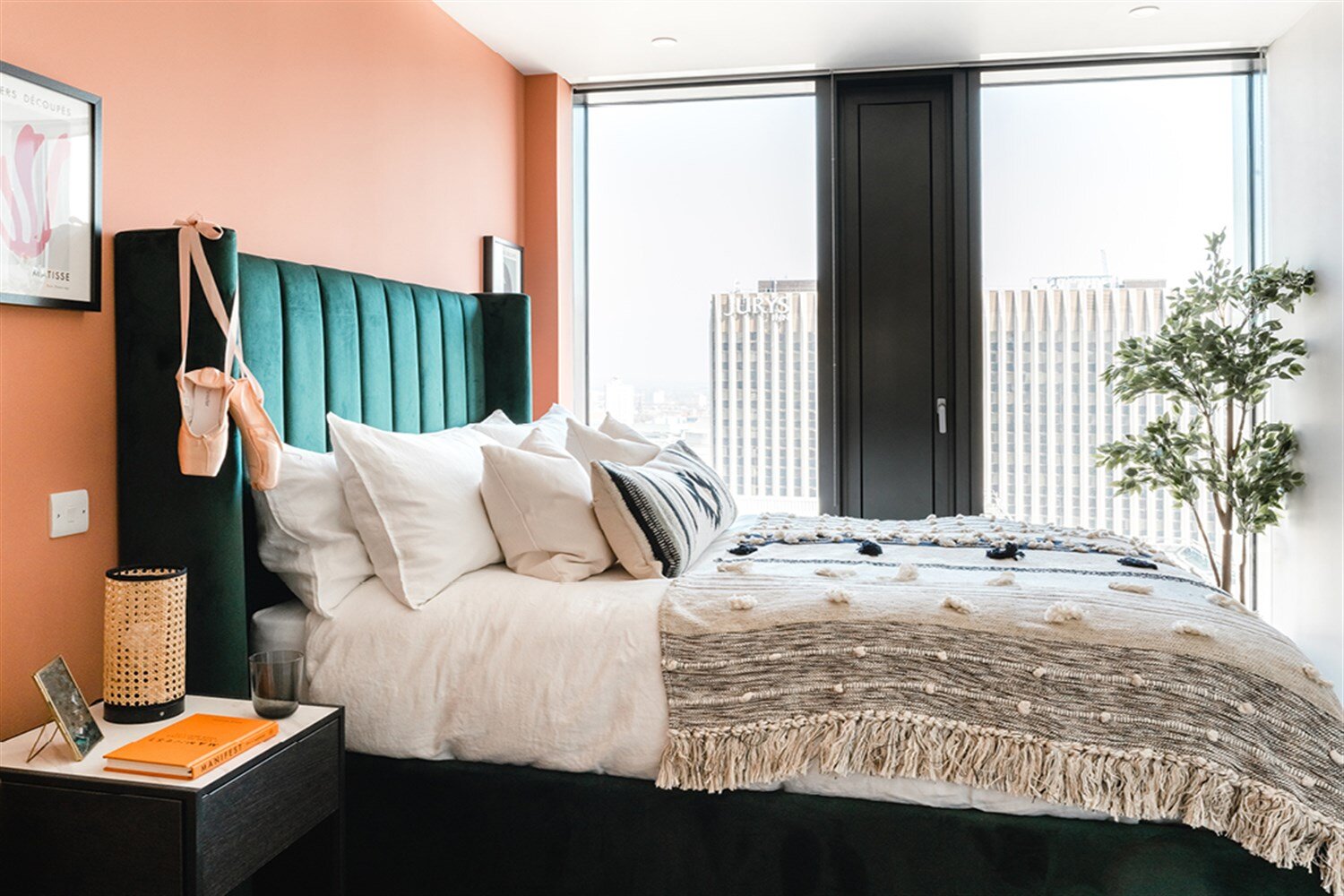 Bedroom with coral walls, a teal velvet headboard and a woven throw beside tall windows