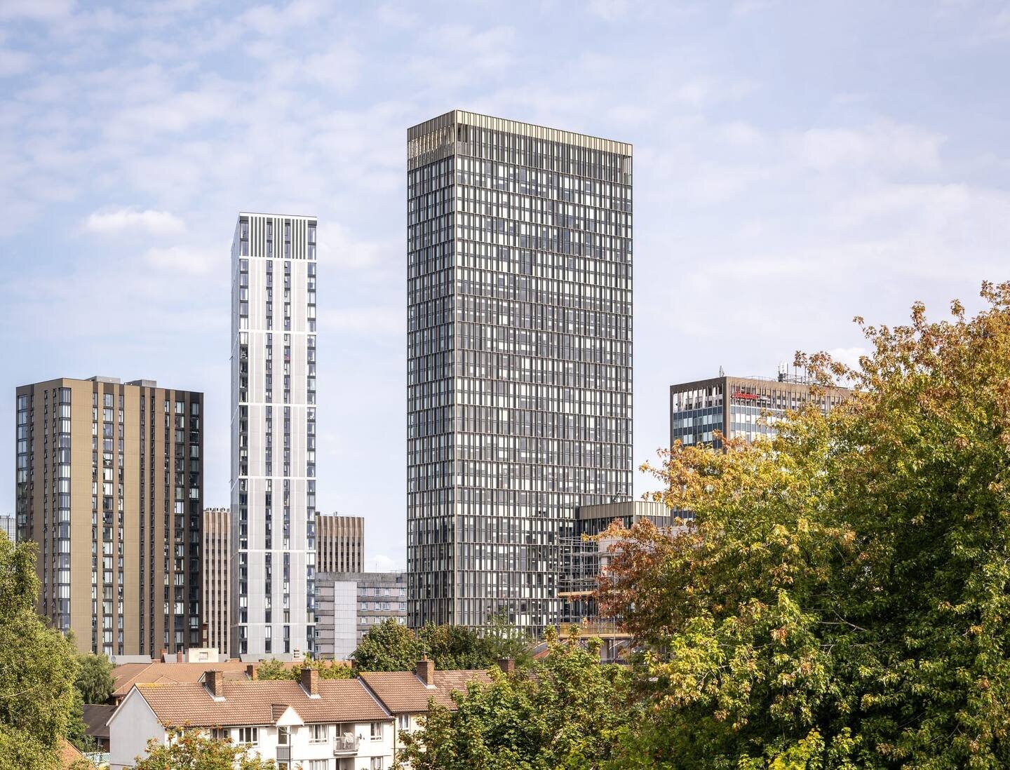The Mercian, a dark tower with a gold crown, rising above trees and rooftops beside a slim white tower
