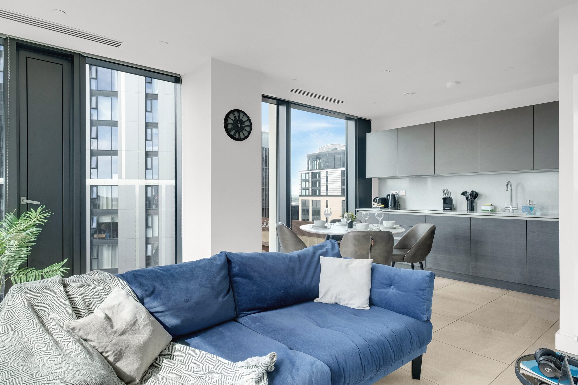 Living room with a blue velvet sofa, the kitchen beyond and tall windows onto the city, at The Mercian, Birmingham